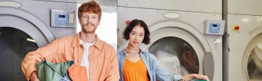 smiling multiethnic couple holding clothes near washing machines in coin laundry, banner clipart