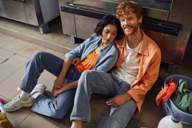 Çamaşırhanede çamaşır makinesinin yanında oturan gülümseyen çok ırklı çiftin yüksek açılı görüntüsü