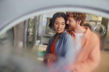 Romantik kızıl saçlı adam çamaşırhanede Asyalı kız arkadaşına sarılıp flört ediyor.