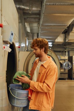 side view of young redhead man holding clothes near washing machine in public laundry clipart
