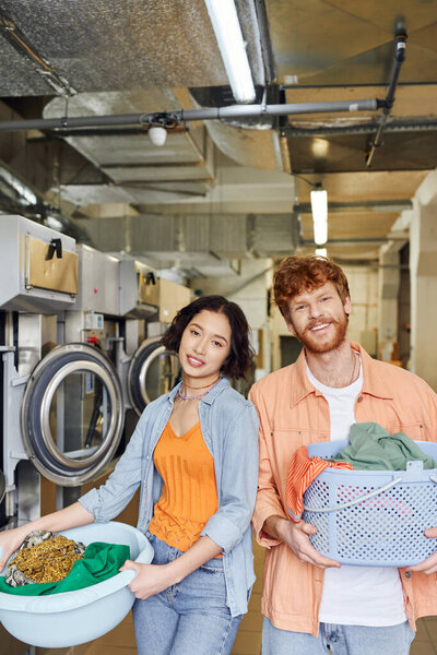 positive interracial couple holding baskets with clothes and looking at camera in coin laundry