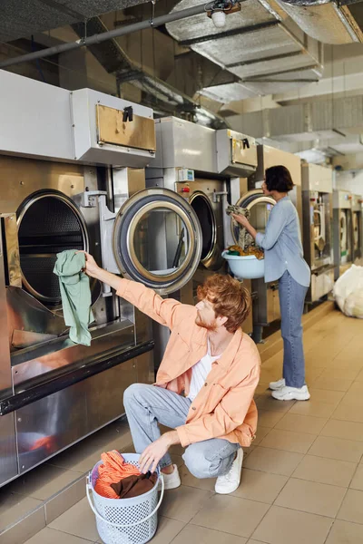 stock image man putting clothes in washing machine near asian girlfriend in public coin laundry