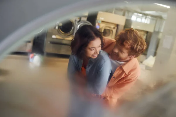 Stock image young multiethnic couple hugging while standing near blurred washing machines in coin laundry