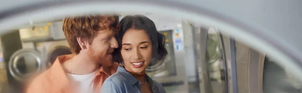 young redhead man whispering to smiling asian girlfriend in blurred public laundry, banner