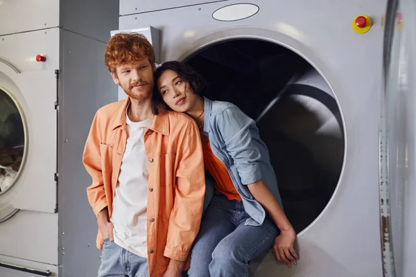 stock image young asian woman sitting on washing machine near redhead boyfriend in public laundry