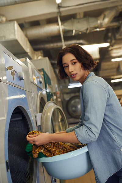 young asian woman looking at camera and holding basket with clothes in public laundry