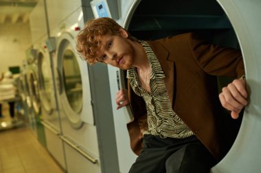 confident young redhead man looking at camera while posing in washing machine in coin laundry clipart