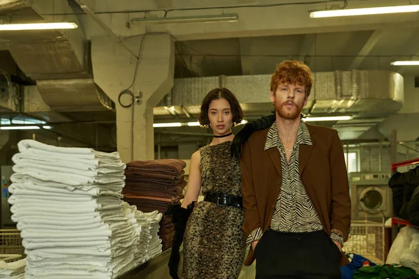 stock image young fashionable interracial couple looking at camera near towels in public laundry