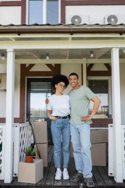 Gülümseyen Afrikalı Amerikalı adam kız arkadaşına sarılıyor ve yeni evin verandasındaki kameraya bakıyor.