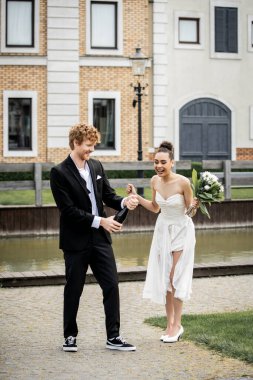 wedding in city, groom opening champagne near excited african american bride with flowers on street clipart