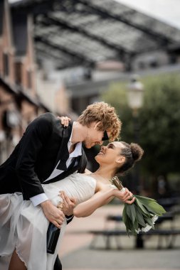 stylish redhead groom embracing african american bride wit bouquet on city street, outdoor wedding clipart