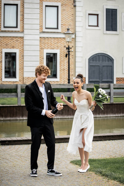 young redhead groom opening champagne near african american bride with bouquet on street