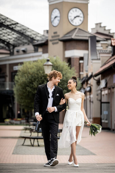 outdoor wedding, young multiethnic couple in elegant attire with champagne and flowers on street