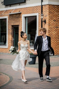 Çok ırklı şık bir çift düğün kıyafetleri içinde el ele tutuşup şehir sokaklarında yürüyorlar.