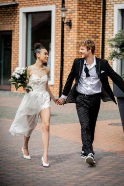 african american bride with flowers and redhead groom in suit walking in city, love outdoors clipart