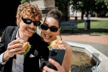 Güneş gözlüklü ırklar arası yeni evliler ellerinde hamburgerler ve şehirdeki akıllı telefonlardan selfie çekiyorlar.
