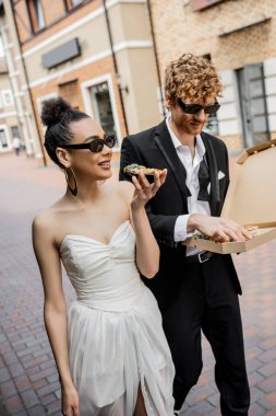 Açık hava kutlaması, şehirde pizzayla yürüyen ırklar arası bir çift, düğün kıyafeti, güneş gözlüğü.