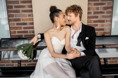 redhead man with flowers sitting face to face with african american bride on bench, outdoor wedding clipart