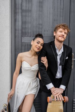 Afro-Amerikan gelini kameraya gülümsüyor. Kızıl saçlı damat binanın, kaykayın ve kaykayın yanında.