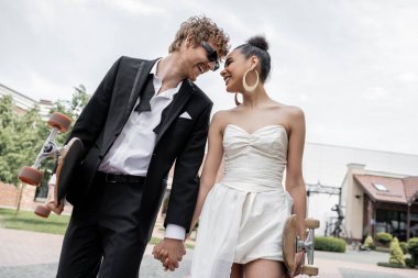 young interracial newlyweds with longboard and skateboard holding hands on street, happiness clipart