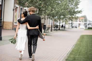 back view of multiethnic elegant newlyweds with bouquet and longboard embracing and walking in city clipart