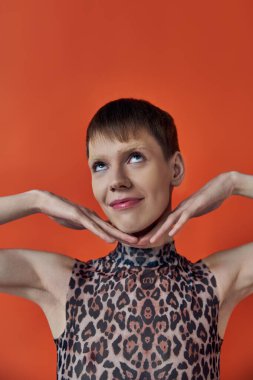 happy androgynous person smiling and posing on orange backdrop, hands near face, queer fashion clipart