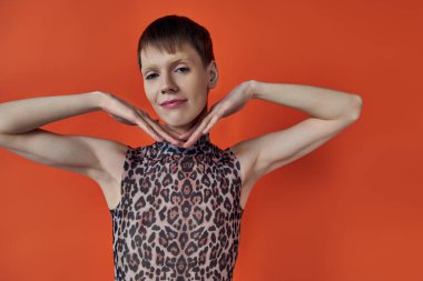 neşeli çift cinsiyetli kişi gülümseyen ve turuncu arka planda poz veren, eller yüze yakın, tuhaf moda