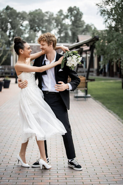 elegant african american bride with bouquet embracing smiling groom on street, joy, urban romance