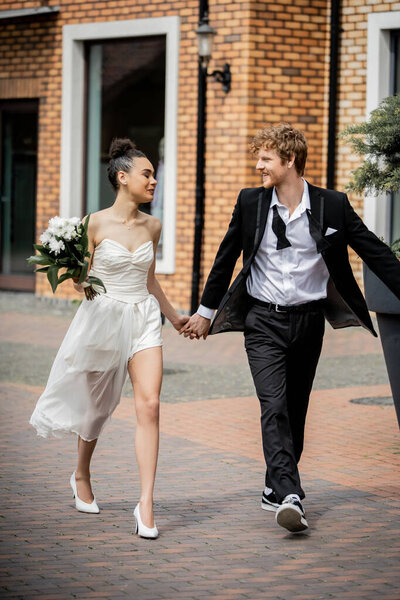 african american bride with flowers and redhead groom in suit walking in city, love outdoors