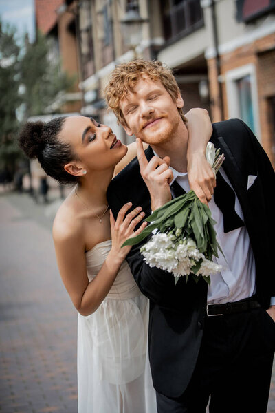 elegant african american bride embracing groom pointing at cheek, wedding celebration in city