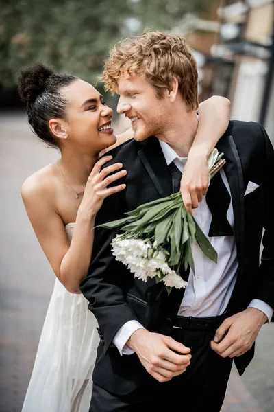 outdoor wedding celebration, excited african american bride with bouquet embracing redhead groom