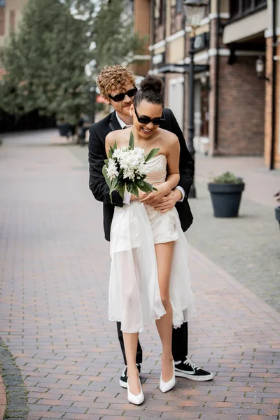 redhead man in sunglasses embracing african american bride with wedding bouquet, wedding in city