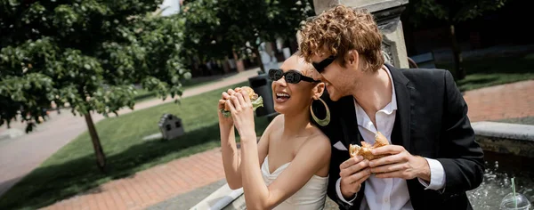 cheerful interracial newlyweds snack with burgers near city fountain, outdoor wedding, banner