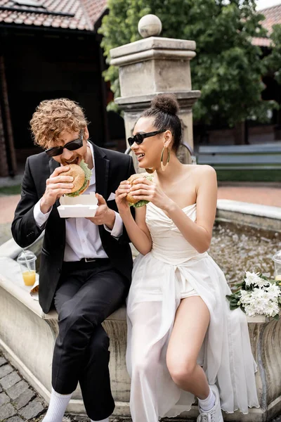redhead man in sunglasses eating burger near african american bride, sitting on city fountain