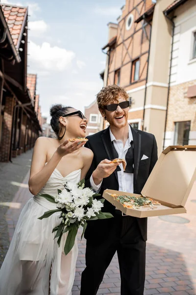 african american bride in sunglasses, with pizza and bouquet, laughing near groom on urban street