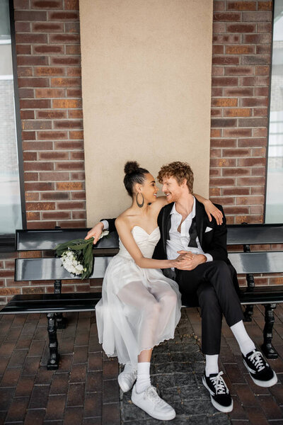 wedding in city, happy redhead groom with bouquet sitting face to face with african american bride