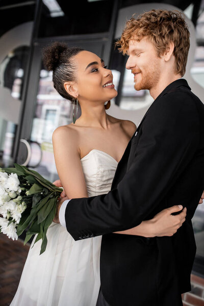 overjoyed and elegant interracial couple in wedding attire embracing on city street