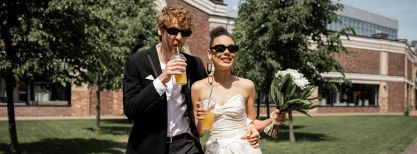 stylish interracial couple in sunglasses, with orange juice and flowers, wedding in city, banner