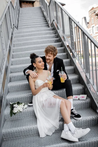 young and happy interracial couple in wedding attire sitting with orange juice on stairs in city