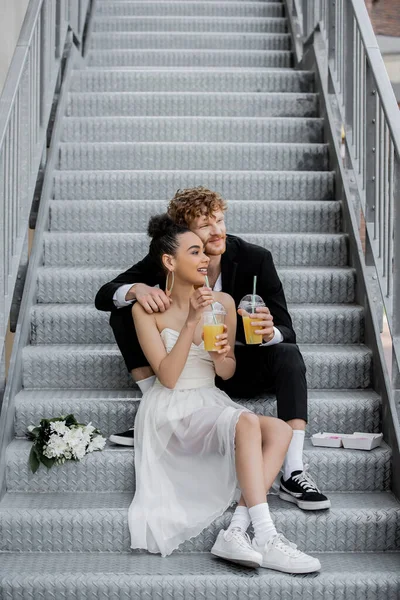 喜庆的跨种族新婚夫妇 橙汁相映成趣 望着街上花束附近的楼梯 — 图库照片