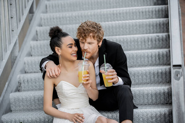 african american bride smiling near redhead groom drinking orange juice on stairs in city