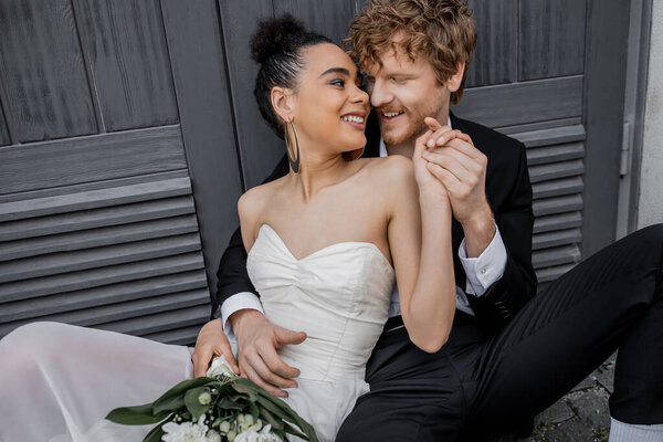 redhead groom holding hands with african american bride sitting with bouquet near city building