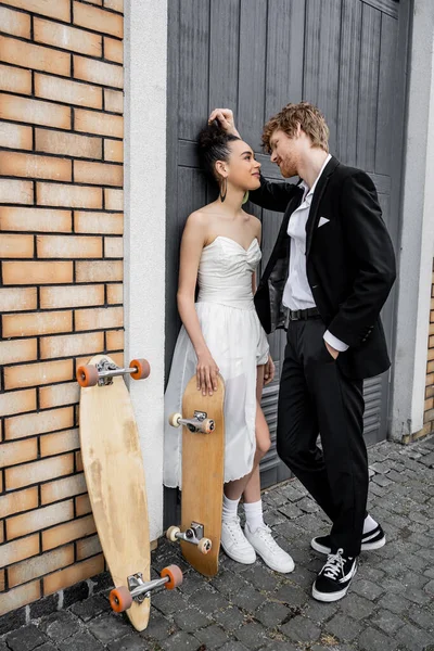 stock image elegant redhead groom with hand in pocket near african american bride, longboard and skateboard