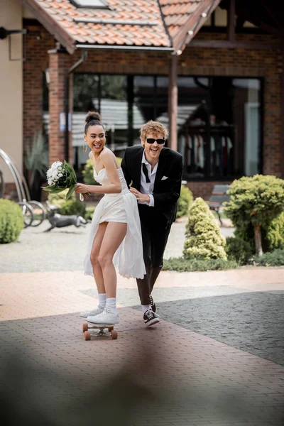 stock image cheerful groom in sunglasses supporting elegant african american bride riding longboard on street