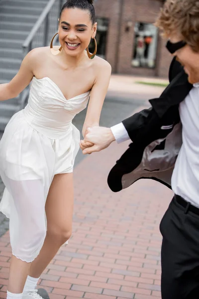 carefree african american woman in wedding dress holding hands with groom and running in city