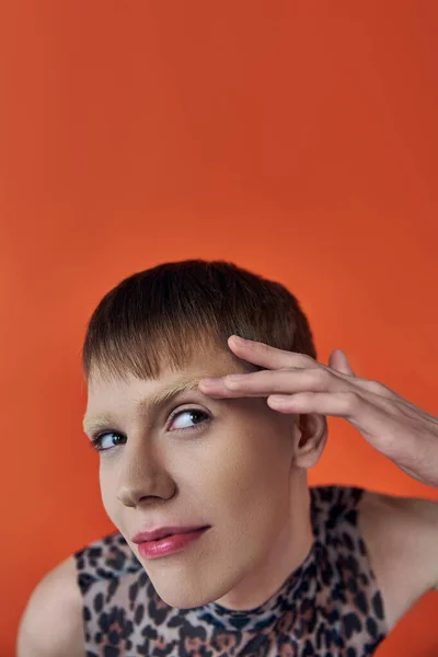 stock image nonbinary person smiling and looking up on orange backdrop, touching eyebrow, queer fashion