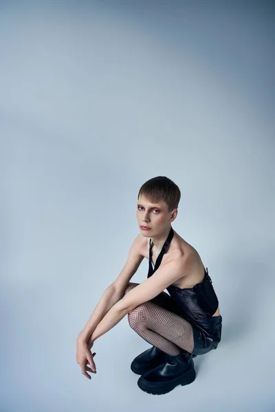 queer model in black corset and shorts sitting on grey backdrop, androgynous person, fashion