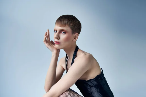 model in corset and fishnet tights sitting on grey backdrop, androgynous person, queer fashion