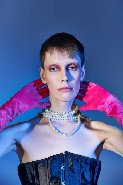 queer person in black corset and pearl necklace posing on blue backdrop, pink gloves, edgy fashion