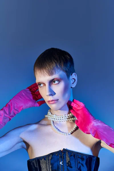 queer person in black corset and pearl necklace posing in pink gloves on blue backdrop, edgy fashion